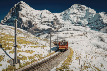 A red cable car travels along a track through a snowy mountain landscape with a clear blue sky.