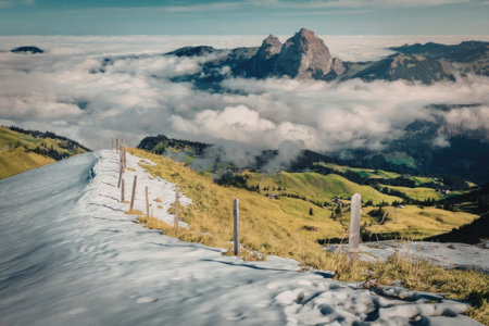 A scenic view of a snow covered mountain peak above the clouds with lush green grass and a fence in...