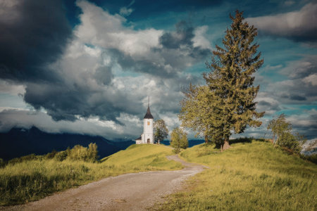 A picturesque scene featuring a small white church on a hill with a dirt road leading to it,...