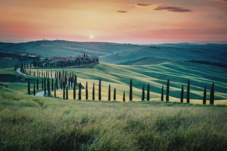 A picturesque landscape of rolling green hills, a winding road, and a fence at sunset.の写真素材