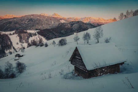 A small wooden cabin sits on a snowy hillside, surrounded by trees and mountains in the background...