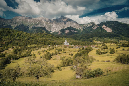 A picturesque landscape featuring a lush green valley with trees and grassy fields, a small white building in the distance, surrounded by majestic mountains under a blue sky with fluffy white clouds.の写真素材