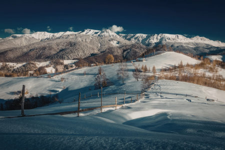 A serene snowy landscape with a fence in the foreground and snow-capped mountains in the background under a clear blue sky.の写真素材