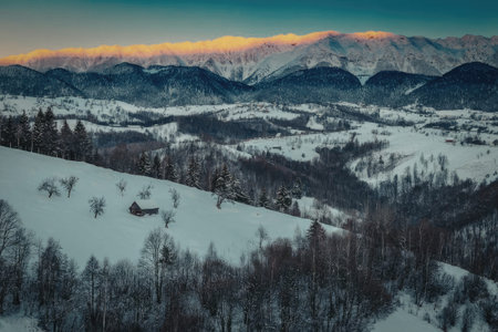 A serene snowy mountain landscape at sunset, with rolling hills and trees covered in snow, set against a backdrop of majestic mountains and a vibrant sky.の写真素材