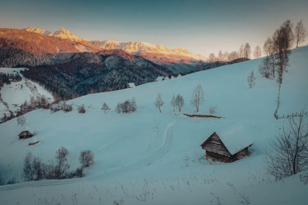A serene snowy mountain landscape at sunset with a small wooden cabin and trees on a hill