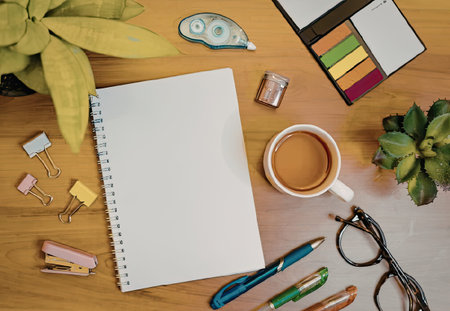 A wooden desk with a spiral notebook, pens, pencils, paper clips, a cup of tea, glasses, and plants.