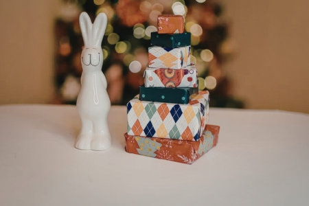 A white rabbit statue stands beside a stack of colorful wrapped Christmas presents on a table with a blurred Christmas tree in the background.の写真素材