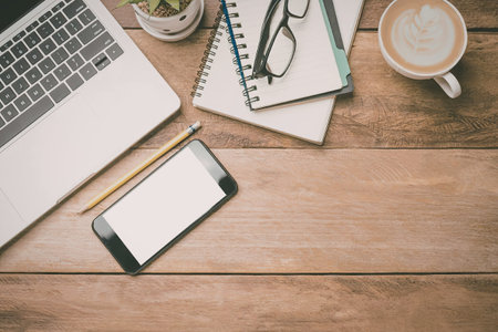 A wooden desk with a laptop, mobile phone, notebooks, glasses, pencil and a cup of coffee
