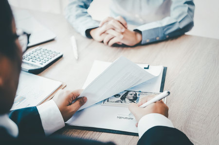 A close-up of a businessman's hands reviewing documents at a desk with another person in the...