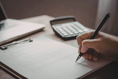A person's hand holding a pen and writing on a piece of paper with a calculator and clipboard in the...