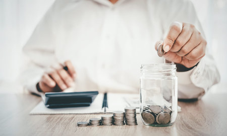 A man in a white shirt sitting at a desk, putting coins into a clear glass jar with some coins...