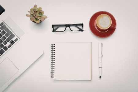 A modern workspace setup with a laptop, notebook, pen, glasses, coffee, and plant on a white desk.