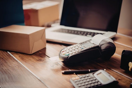 A cluttered office desk with a laptop, payment terminal, calculator, pen, and cardboard boxes.