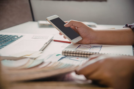 A person is sitting at a desk, holding a phone and surrounded by a notebook, papers, and a laptop.