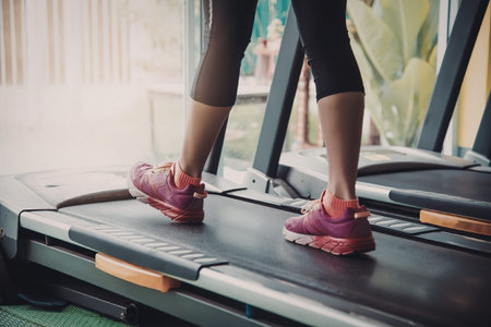 A woman wearing pink sneakers and black leggings runs on a treadmill in a gym with a blurred...