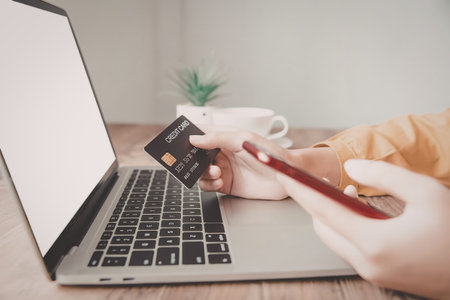 A person is sitting at a desk with a laptop, credit card, and smartphone, likely making an online...