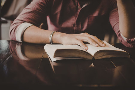 A person wearing a pink shirt and a bracelet sits at a table reading an open book.