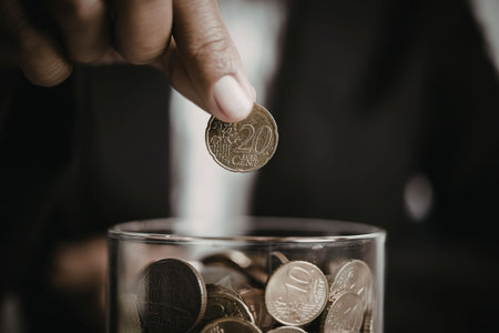 A person's hand putting a coin into a glass jar filled with coins, symbolizing saving or investment.