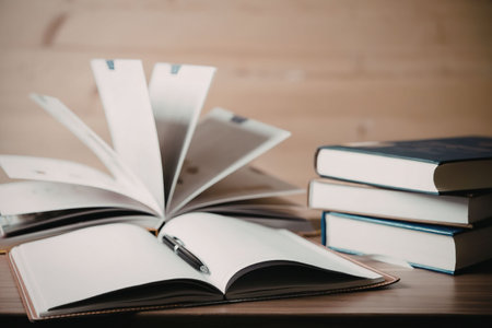 An open book lies on a wooden table with its pages fanned out, accompanied by a stack of closed...