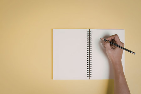 A hand holding a pencil over a blank spiral-bound notebook on a yellow background.