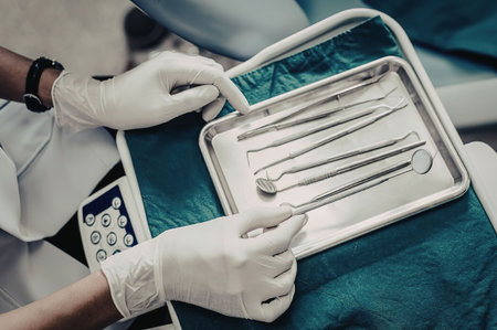 A gloved hand holding dental tools over a tray with other tools, in a medical environment.