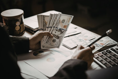 A person is sitting at a desk, holding a stack of US dollar bills in one hand and a pen in the...