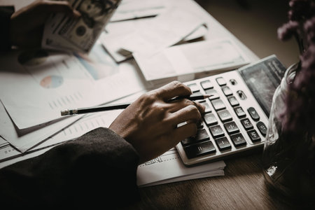 A person is sitting at a desk, using a calculator and surrounded by papers and money.