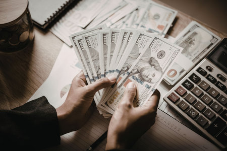 A person holding a fan of US dollar bills over a desk with a calculator, papers, and more cash.