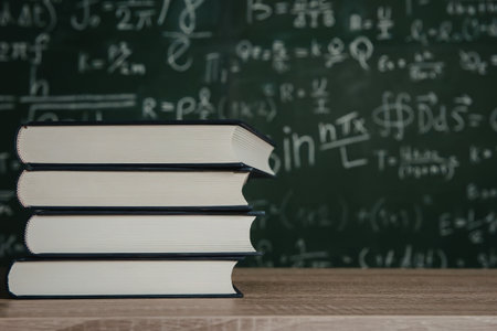A stack of four books with white covers and black spines sits on a wooden desk in front of a green...