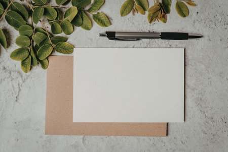 A blank white card on top of a brown envelope with a black pen and green leaves on a marble surface