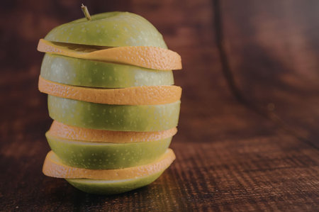 A green apple with slices of orange wrapped around it, sitting on a dark wooden surface.