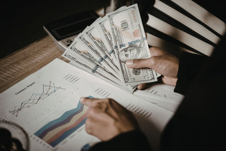 A person holding a stack of US dollar bills and a financial document with graphs and charts.