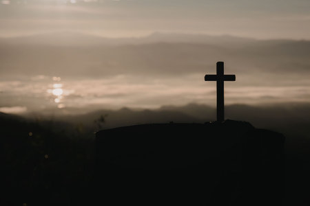 A silhouette of a cross on top of a hill during sunset, with clouds and sun in the background.