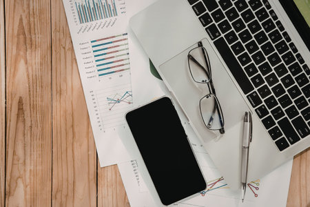 A laptop, smartphone, glasses, and pen on a wooden desk with papers and charts.