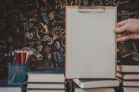 A clipboard with a blank white paper on it, placed on a desk with books, a pencil holder, and a hand...
