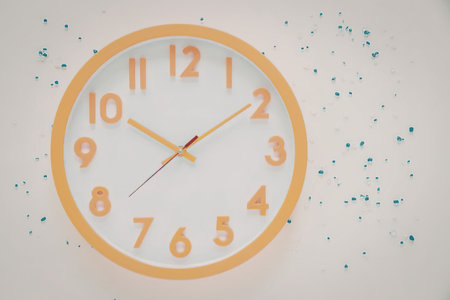 An orange clock with a white face is mounted on a white wall, surrounded by blue and green speckles.