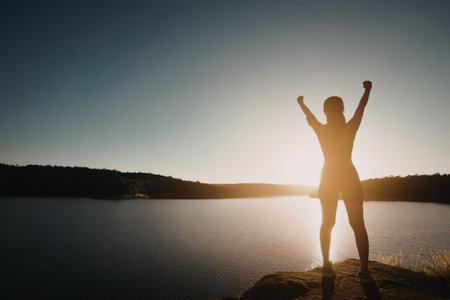 A woman stands on a rock with her arms raised in triumph as the sun sets behind her over a serene...