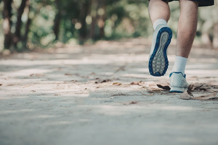 A person's legs and feet as they run on a path covered with leaves and surrounded by trees.