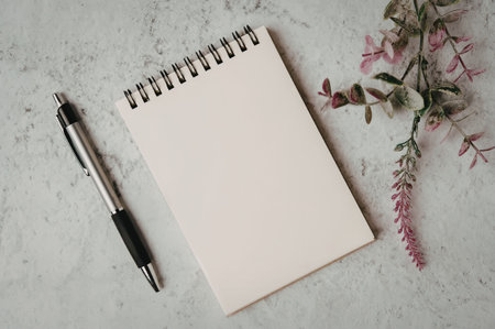 A spiral-bound notepad and pen placed on a marble surface with pink flowers