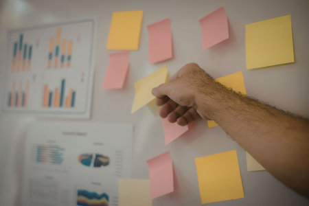A person's hand placing a pink sticky note on a wall with various colorful sticky notes and charts.