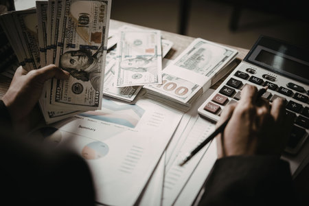 A person is sitting at a desk, calculating money with a calculator and holding bills.
