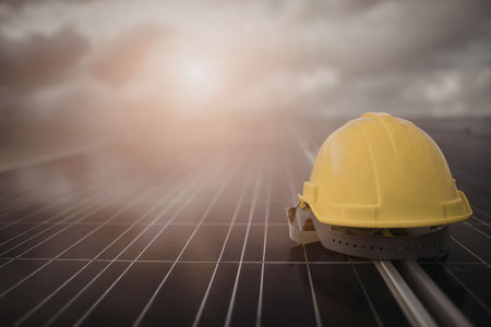 A yellow hard hat sits on a solar panel on a cloudy day with the sun shining through the clouds.