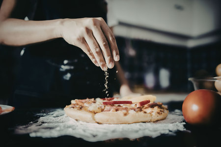 A person is seasoning a pizza in a kitchen. The pizza has a thick crust and is topped with red peppers and cheese. A tomato and a bowl of ingredients are on the counter.の写真素材