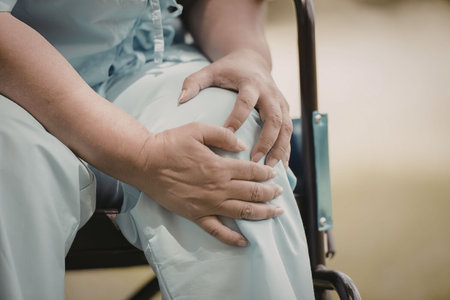 A close-up of an elderly person sitting in a wheelchair, clutching their knee in discomfort,...