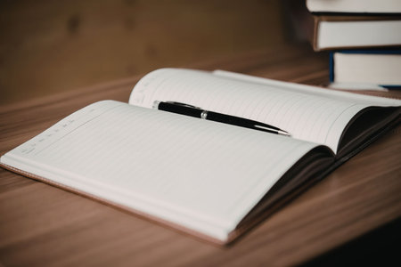 An open book with blank pages lies on a wooden desk, accompanied by a stack of books in the...