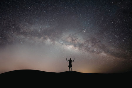 A silhouette of a man standing on a hill with arms raised under a starry night sky with the Milky...