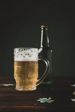 A glass of beer with foam and a bottle of beer on a dark wooden table with shamrocks scattered...