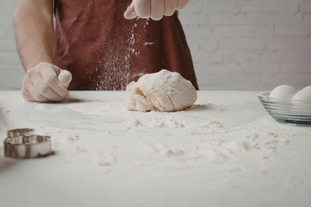 A person wearing a red apron is kneading dough on a floured surface with a bowl of eggs and cookie...