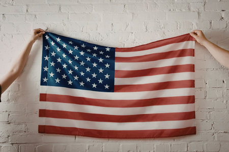 A person holding an American flag against a white brick wall, showcasing the flag's vibrant red,...