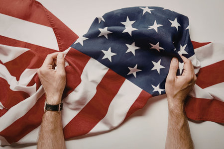 A close-up of two hands holding an American flag against a white background, with the flag's stars...
